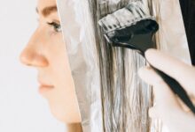 A woman carefully sectioning her hair for at-home highlights, with foils, a tint brush, gloves, and hair-lightening products arranged beside her.