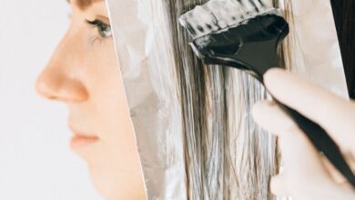 A woman carefully sectioning her hair for at-home highlights, with foils, a tint brush, gloves, and hair-lightening products arranged beside her.