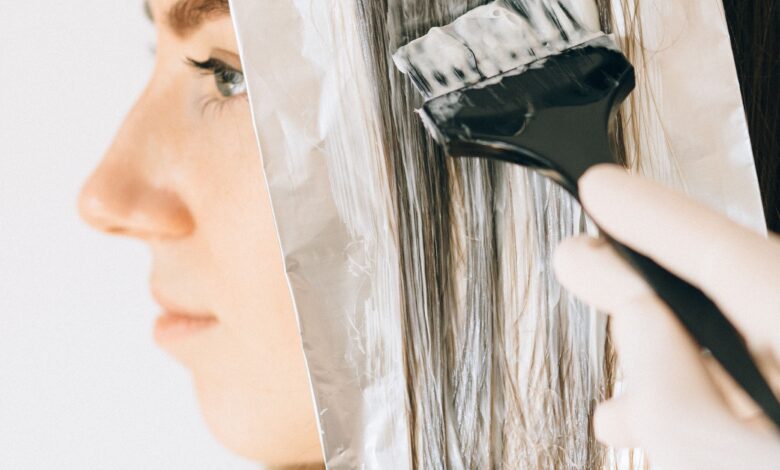 A woman carefully sectioning her hair for at-home highlights, with foils, a tint brush, gloves, and hair-lightening products arranged beside her.