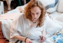 Fashion designer working at a studio desk with sketches, fabric swatches, measuring tools, and clothing samples.